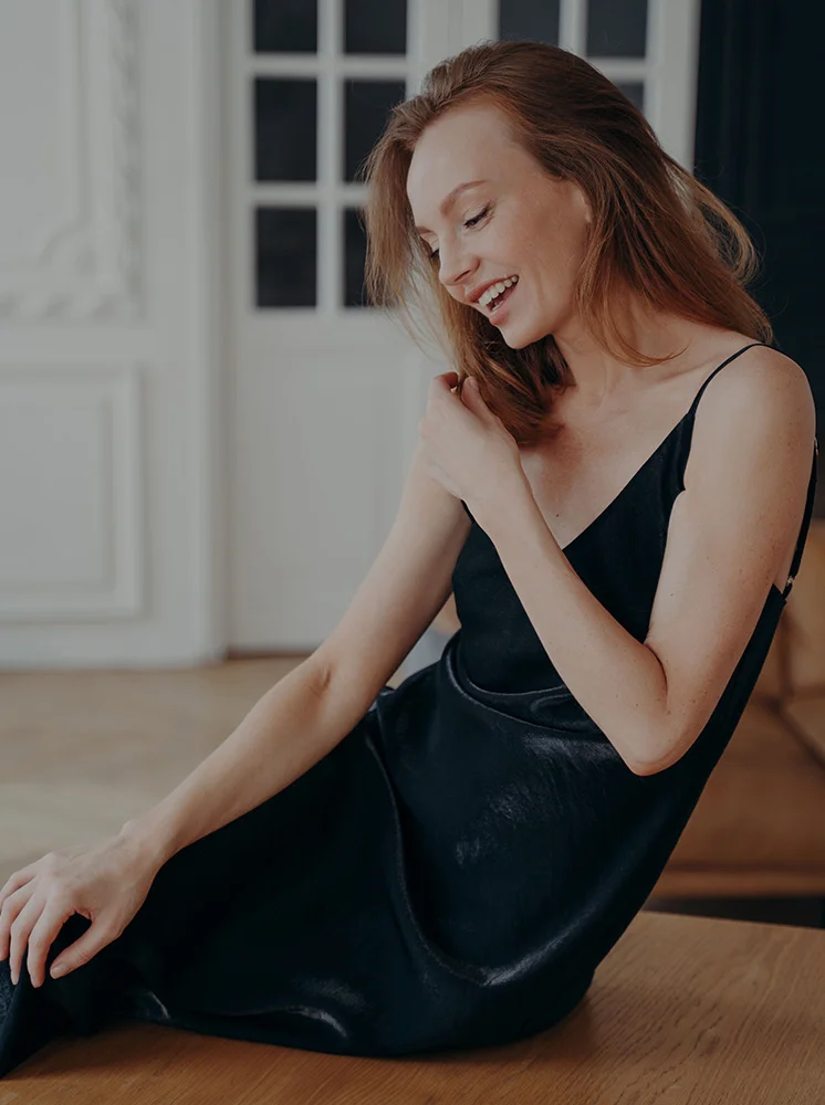 A light-skinned woman with shoulder-length reddish-brown hair sits on a light-colored wooden floor. She wears a black, sleeveless, midi-length dress with a silky texture. Looking to her right with a slight smile, her posture is relaxed, and her hands rest on the dress. The background features a light-colored wall with a white door and window panes, and soft, diffused lighting. - Arm Lift Surgery in New York and New Jersey