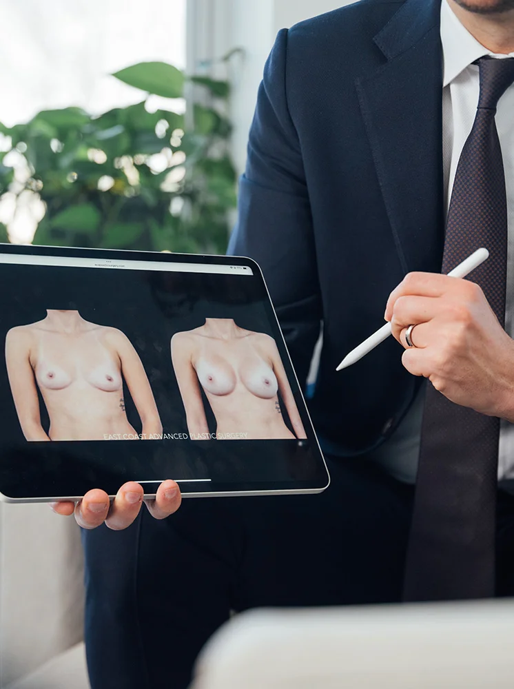 A person in a dark-navy blue suit and a dark-purple tie holds a tablet displaying before-and-after images of a woman's breasts, likely showing the results of a cosmetic procedure. The person holds the tablet with one hand while using a stylus or pen with the other. The background includes part of a plant and a blurred light-colored wall and window. - Breast Augmentation in New York and New Jersey