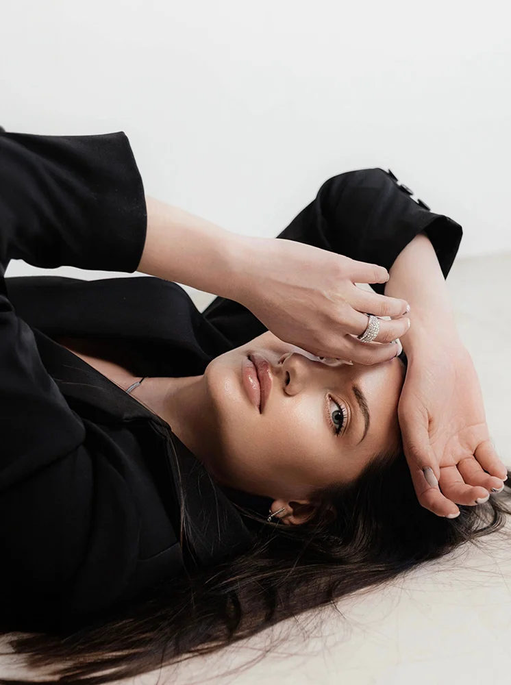 A young woman with long, dark brown hair lies on a light beige surface, wearing a black blazer. Her arms rest on her forehead, and she gazes downward and slightly left. With light skin, soft pink lips, and neutral makeup, she wears a small silver necklace and several rings. The soft, diffused lighting creates a calm, stylish atmosphere, with a plain light cream background drawing focus to her. - Brow Lift in New York and New Jersey