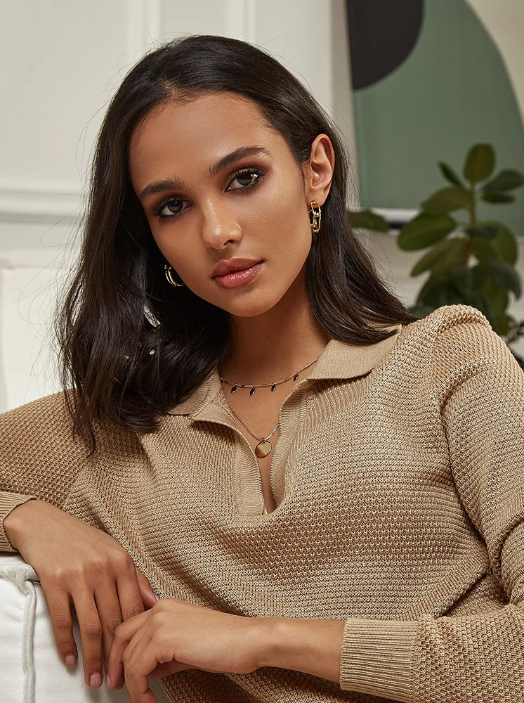 A woman with dark brown hair and a warm, beige complexion is seated, looking at the camera. She wears a light beige, knitted, polo-style sweater and gold hoop earrings. The background features a muted, neutral-colored wall and blurred foliage. Her arms and hands are visible in the foreground. - Dermal Fillers in New York and New Jersey