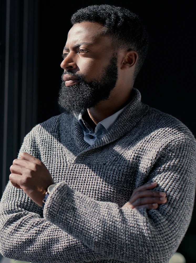 A man of African descent is seen from the mid-chest up, with short dark hair and a thick beard. He wears a light gray, textured sweater with a shawl or mock turtleneck collar and has his arms crossed. His gaze is directed to the right. The background is a blurred dark space, possibly a window or doorway. A wristwatch is visible on his left wrist. The lighting suggests sunlight or bright indoor light, casting subtle shadows. - Excisional Procedures in New York and New Jersey
