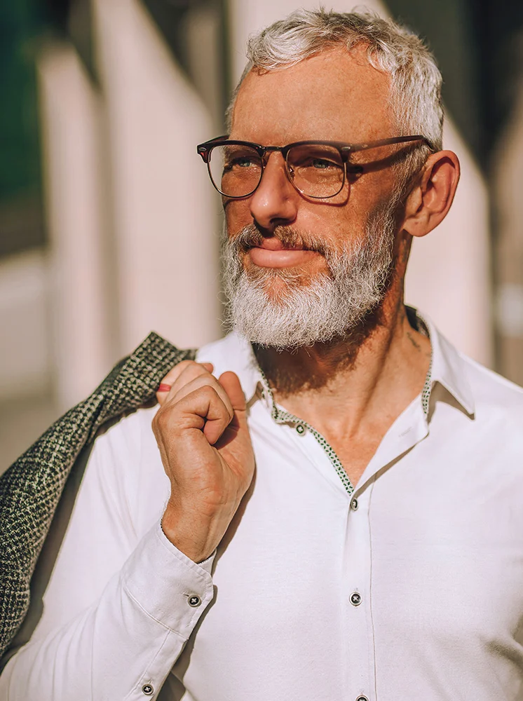 A close-up of a middle-aged man with short, graying hair and a full gray beard. He wears glasses with dark brown frames, a white button-down shirt with subtle green or dark gray detailing, and a dark, patterned jacket draped over his shoulder. His expression is neutral and contemplative, with natural light highlighting his face. The background is out of focus, suggesting a light beige or cream-colored architectural structure. - Eyelid Surgery for Men in New York and New Jersey