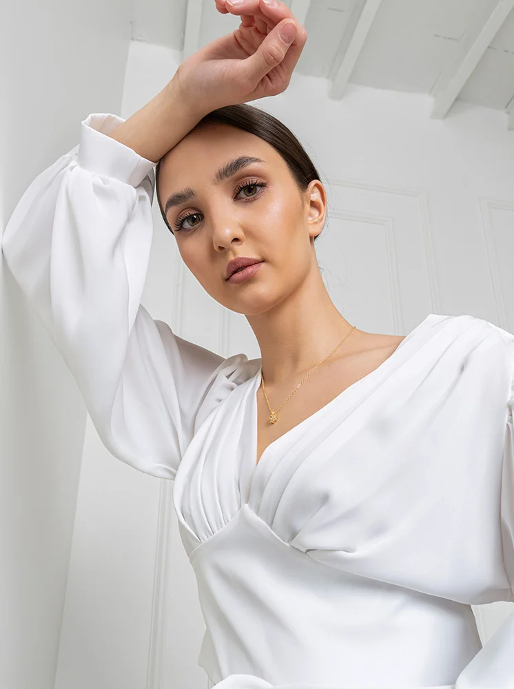 A woman is seen from a low-angle, close-up perspective. She has light skin and shoulder-length dark brown hair styled in a bun. Her makeup is natural, with eyeliner, mascara, and subtle blush and bronzer. Her expression is neutral and thoughtful. She wears a white, V-neck, wrap-style top with full puffed long sleeves, featuring pleats and a small gold pendant on a delicate chain. The background is a plain white wall with a light-colored textured ceiling, and soft lighting enhances the elegant, modern aesthetic. - Eyelid Surgery in New York and New Jersey