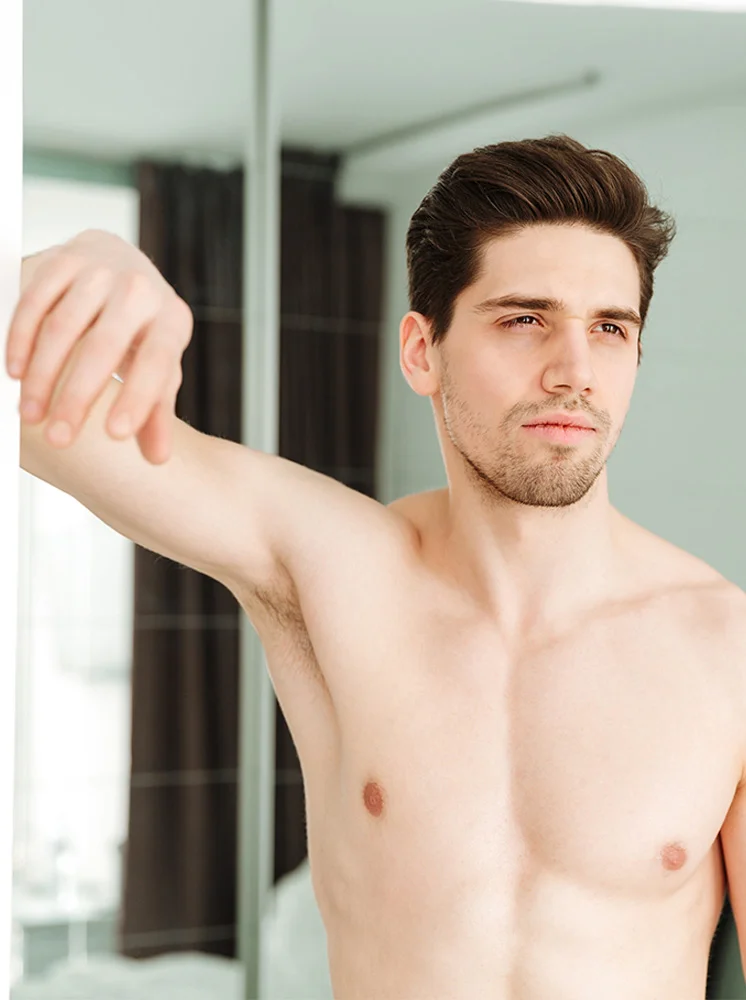 A light-skinned man with short, dark brown hair and a short trimmed beard is seen from the mid-chest up, shirtless. His arm is bent at the elbow, resting near the side of the wall, while his gaze is directed to the right. The background is blurred, featuring a reflection of a room with hints of brown curtains and a light beige or off-white wall. - Female to Male Top Surgery in New York and New Jersey