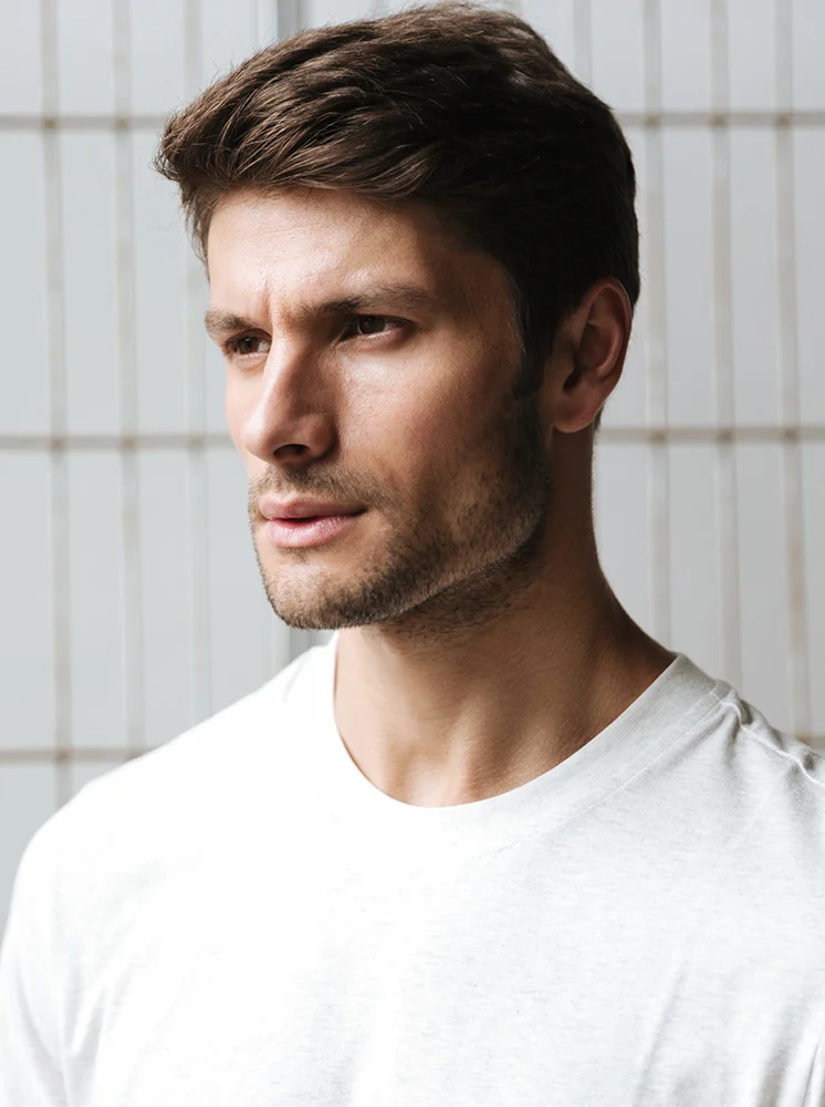 A close-up of a man’s head and upper torso. He has short, dark brown hair, a light complexion, and a short, trimmed beard. His expression is neutral, and he’s looking slightly off to his right. He’s wearing a plain, light beige or white round-neck T-shirt. The background is a light gray, patterned wall with vertical lines. - Injectables for Men in New York and New Jersey