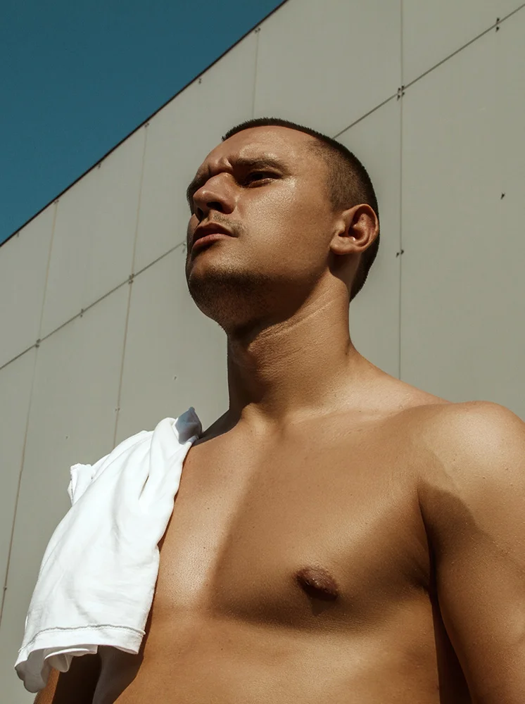 A man with a short dark haircut and light tan complexion is seen from the mid-chest up, outdoors. He gazes upward to his right with a white t-shirt draped over his shoulder. Sunlight casts strong shadows across his chest. A beige-gray paneled wall and clear blue sky form the background. - Laser Hair Removal for Men in New York and New Jersey