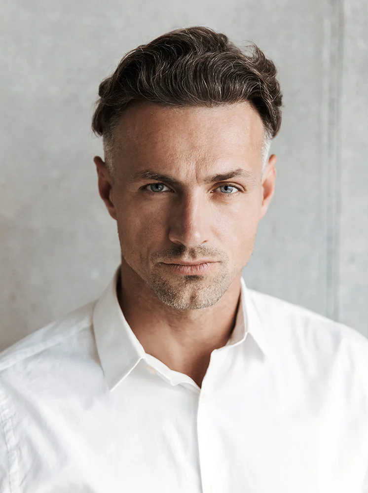 Close-up portrait of a light-skinned man with short, dark brown and gray hair styled in a slightly wavy, tousled look. He has a neutral, serious expression and light blue eyes, gazing directly at the camera. He is wearing a crisp white button-down shirt with a smooth texture. The background is a light gray concrete wall, and the even lighting highlights his features clearly without harsh shadows. - Lumecca in New York and New Jersey