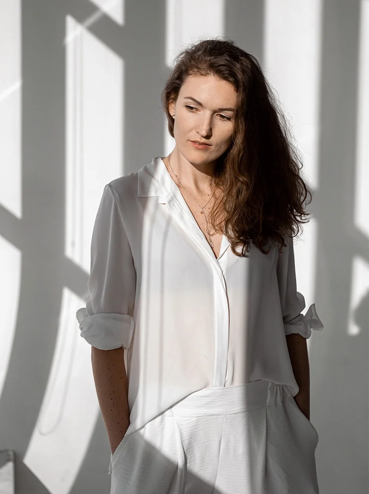 A woman with shoulder-length brown hair stands in front of a white wall with vertical shadows. She wears a flowing, white, long-sleeved button-down shirt and matching white pants made of smooth, slightly textured fabric. Her gaze is downward and slightly to the left, with a pensive expression. Bright lighting creates soft shadows along her body, enhancing the dramatic effect. A delicate gold chain necklace is visible. The plain white background focuses attention on the woman and her attire. - Lymphovenous Bypass LVB in New York and New Jersey