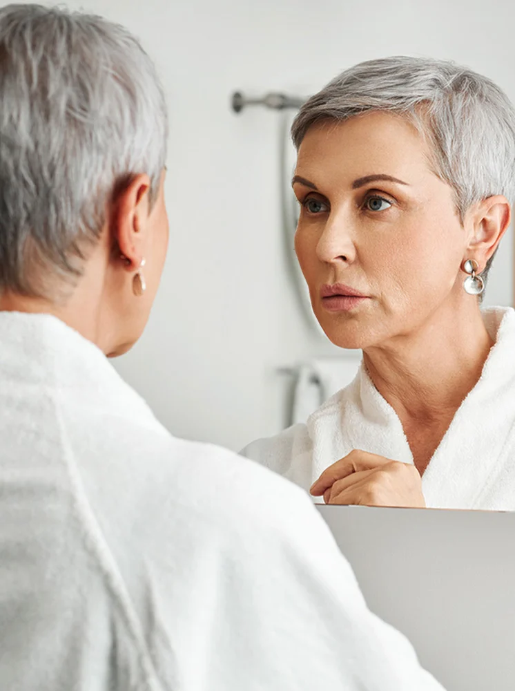 A woman with short gray hair is looking at her reflection in a bathroom mirror, wearing a white bathrobe. Her reflection shows a woman of similar age, with short gray hair and visible wrinkles on her face and neck. The woman in the robe gazes directly at her reflection, her expression contemplative or concerned, as if inspecting her appearance. The background features light, neutral bathroom colors. The mirror reflects both the woman and the slightly out-of-focus bathroom environment. The focus is on the woman and her reflection. - Minimally Invasive Tissue Excision MITE in New York and New Jersey