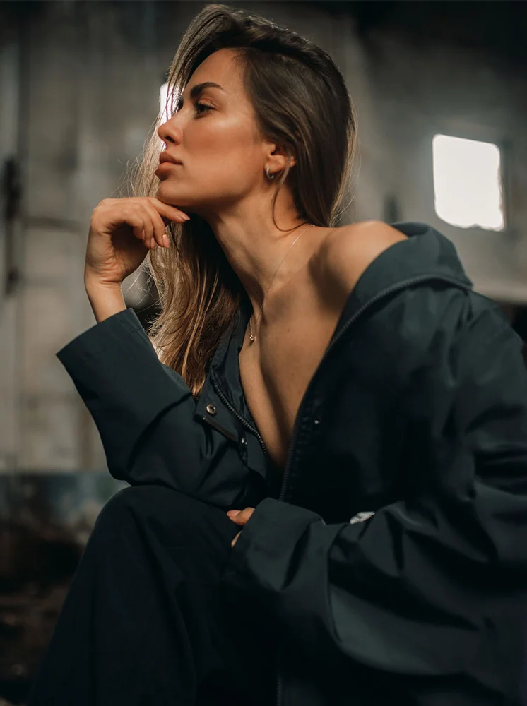 A woman with shoulder-length brown hair sits in a dark jacket, her bare shoulders visible. She gazes to the side, hand resting on her chin. The background is a muted gray, with soft, ambient lighting highlighting her face. - Necklift in New York and New Jersey