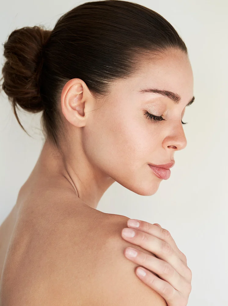 A close-up profile of a woman with light-toned skin and dark brown hair in a bun. Her eyes are closed, and she has a serene expression. She gently rests her hand on her shoulder, with her upper back visible. The background is a soft off-white, creating a calm, natural aesthetic. Her lips are light pink, and her nails are a nude shade. - Nipple Areola Tattooing after Breast Reconstruction in New York and New Jersey