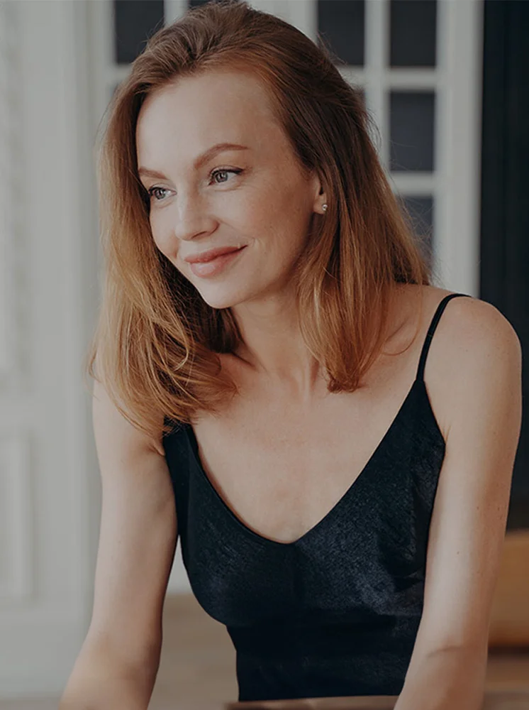 A woman with shoulder-length reddish-brown hair is seated, smiling gently as she looks to her right. She wears a black sleeveless v-neck top. The soft lighting and out-of-focus background, with a white wall and window panes, create a serene atmosphere. - Oncoplastic Reconstruction in New York and New Jersey