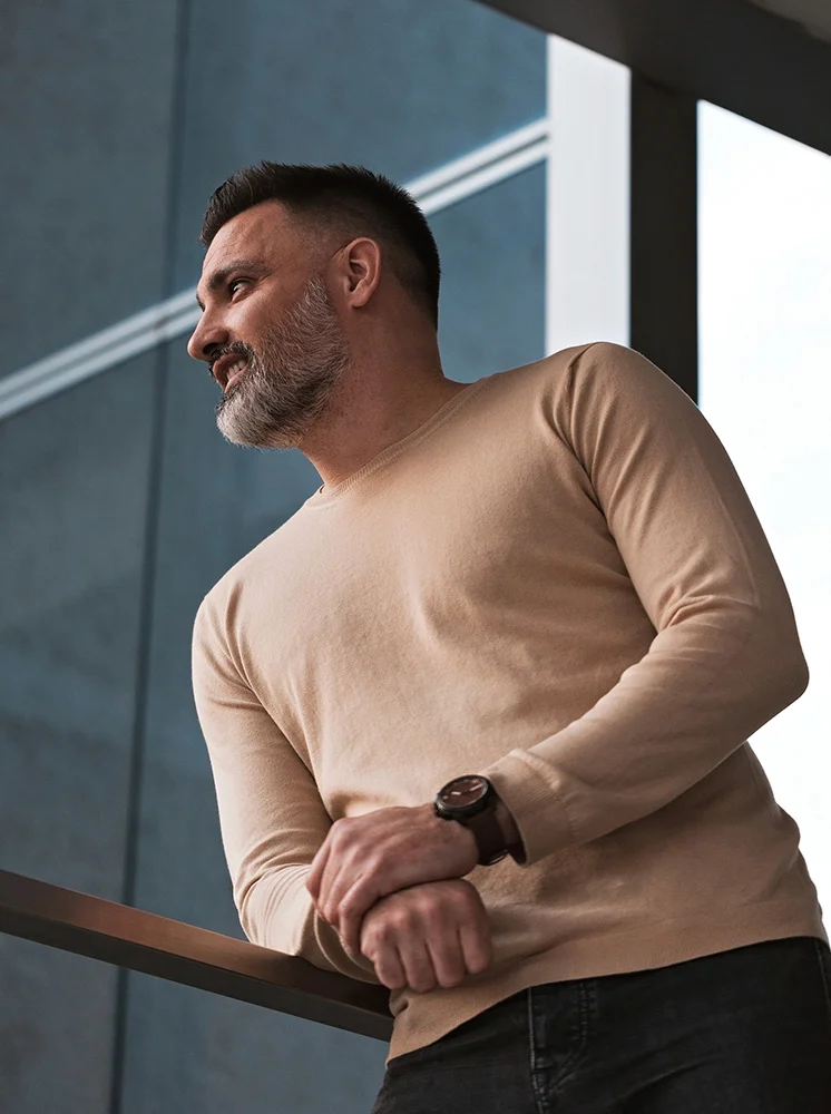 A man with short dark hair and a gray beard leans against a dark metal railing, seen from the mid-chest up. He wears a light beige, long-sleeved sweater and a dark brown watch on his right wrist. His gaze is directed upward and to his right, with a thoughtful expression. The background features a modern, dark-gray building facade with structural elements visible, adding an urban atmosphere to the scene. - Preventative Lymphatic Surgery in New York and New Jersey