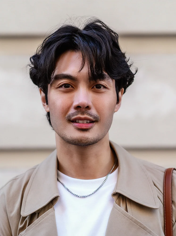 Close-up of a light-skinned man with short, tousled dark hair and a neatly trimmed beard. He wears a beige trench coat over a white t-shirt and a thin silver chain necklace. His expression is neutral, with slightly parted lips. The background is a light beige wall, with a brown leather bag strap visible on the right. - Rhinoplasty for Men in New York and New Jersey