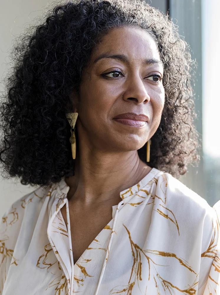 A medium close-up of a middle-aged woman with dark, curly hair and a light brown complexion. She wears a cream blouse with a subtle golden floral print and gold pointy earrings. Her expression is thoughtful, gaze slightly downward. Soft window light gently illuminates her face, with a blurred indoor background. - Vascularized Lymph Node Transfer VLNT in New York and New Jersey