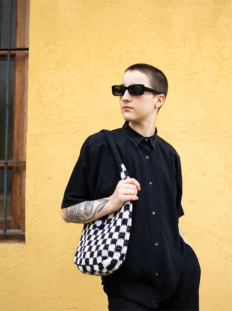 transgender person posing with sunglasses in front of a yellow wall with a black and white checkered purse and black shirt