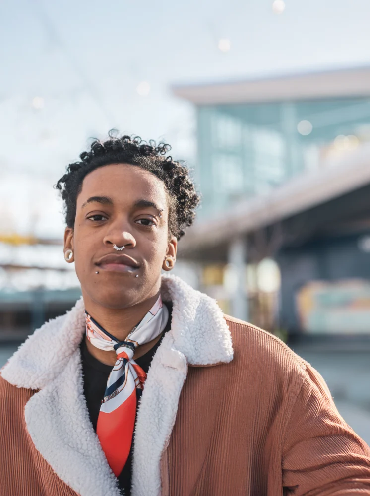 gender fluid person with curly hair, a nose piercing, gauges and neck scarf posing for camera